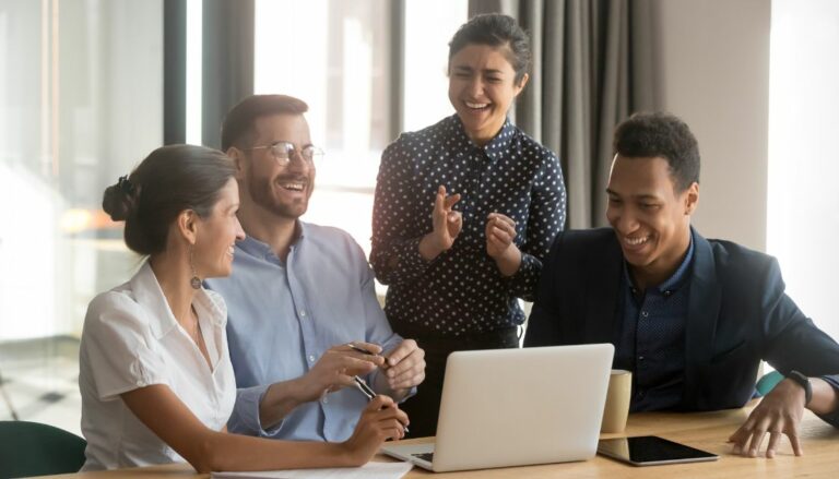 Eine Gruppe von vier Kollegen sitzt an einem Tisch und lacht gemeinsam, während sie auf einen Laptop-Bildschirm schauen. Die lockere und humorvolle Stimmung zeigt, wie Humor am Arbeitsplatz das Team verbindet und die Zusammenarbeit erleichtert. Solche humorvollen Momente während Meetings fördern das Vertrauen unter den Kollegen und machen den Arbeitsalltag angenehmer.