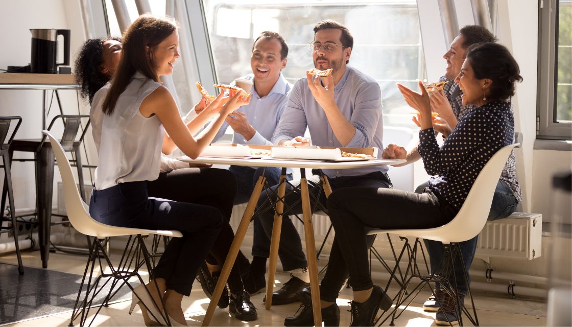 Eine Gruppe von sechs Kollegen sitzt gemeinsam an einem Tisch und genießt eine entspannte Pizza-Pause. Alle lachen und plaudern miteinander, was den Effekt von Humor am Arbeitsplatz auf das Teamgefühl und die Motivation unterstreicht. Solche ungezwungenen Pausen mit einer Prise Humor stärken den Zusammenhalt und sorgen für ein positives Arbeitsklima.