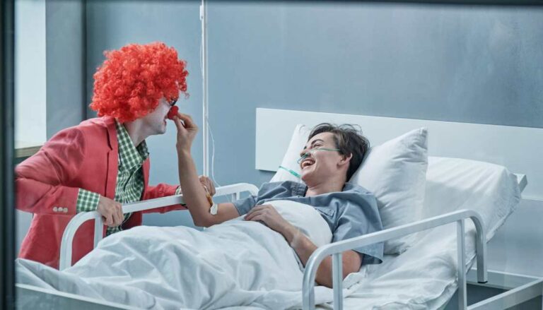 Ein Clown-Arzt mit auffälliger roter Perücke, roter Nase und farbenfroher Kleidung beugt sich zu einer Patientin im Krankenhausbett, die lächelnd eine rote Clownsnase hält. Beide lachen herzlich und schaffen einen warmen, freudigen Moment mitten in der klinischen Umgebung. Dieses Bild zeigt eindrucksvoll, wie Humor hilft heilen, indem er selbst in ernsten Situationen Leichtigkeit und Freude bringt und den Heilungsprozess unterstützt.