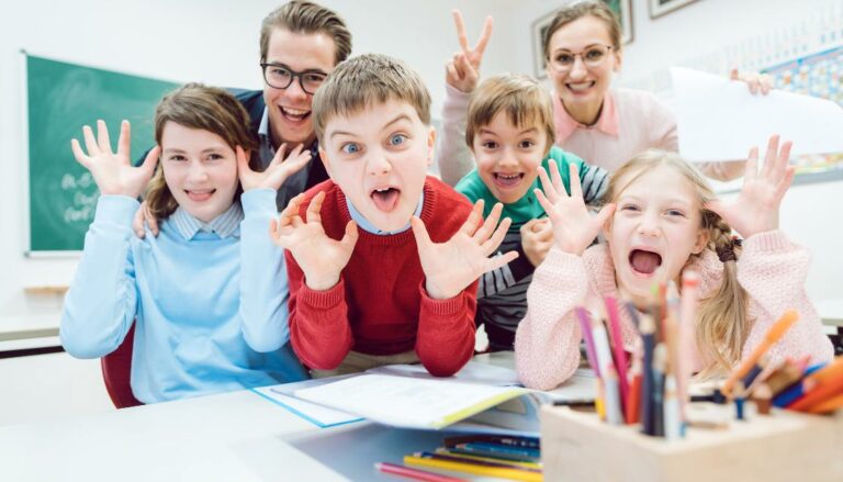 Vier Kinder und zwei Lehrer posieren mit lustigen Grimassen für ein Foto im Klassenzimmer und zeigen so Humor im Unterricht. Die Kinder strecken die Zungen heraus oder machen „Hasenohren“-Gesten, während die Lehrer lächelnd mitmachen. Auf dem Tisch liegen bunte Stifte und Hefte, was auf kreative Aufgaben hinweist, die in einer spielerischen Weise bearbeitet werden. Diese Szene zeigt, wie Humor im Unterricht die Kreativität der Schüler fördert und ihnen ermöglicht, sich frei auszudrücken.