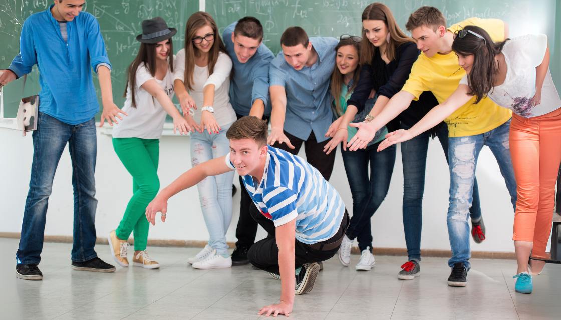 Ein Teenager-Junge in einem gestreiften T-Shirt posiert lachend in einer Breakdance-Haltung am Boden, während etwa acht weitere Teenager ihn umringen und begeistert auf ihn zeigen – ein dynamisches Beispiel für Humor im Unterricht. Die Jugendlichen, lässig gekleidet, lachen ausgelassen und zeigen mit ausgestreckten Armen auf ihn. Diese humorvolle Szene im Klassenzimmer zeigt, wie humorvolle und kreative Ausdrucksformen die Gruppendynamik stärken und eine lockere Lernatmosphäre fördern, in der die Schüler aktiv und spielerisch beteiligt sind.