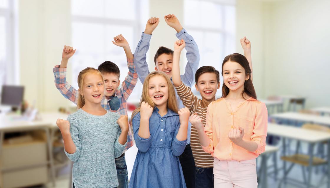 Sieben Grundschulkinder stehen jubelnd im Klassenzimmer und recken voller Freude die Fäuste in die Luft, was Humor im Unterricht aufgreift. Die Kinder, in bunten Outfits gekleidet, lächeln stolz und strahlen ein starkes Gemeinschaftsgefühl aus. Der helle Raum im Hintergrund mit großen Fenstern verstärkt die positive Stimmung. Diese Szene vermittelt Freude am Lernen und zeigt, wie Humor im Unterricht das Selbstbewusstsein und die Motivation der Schüler steigern kann, indem Erfolgserlebnisse gemeinsam gefeiert werden.