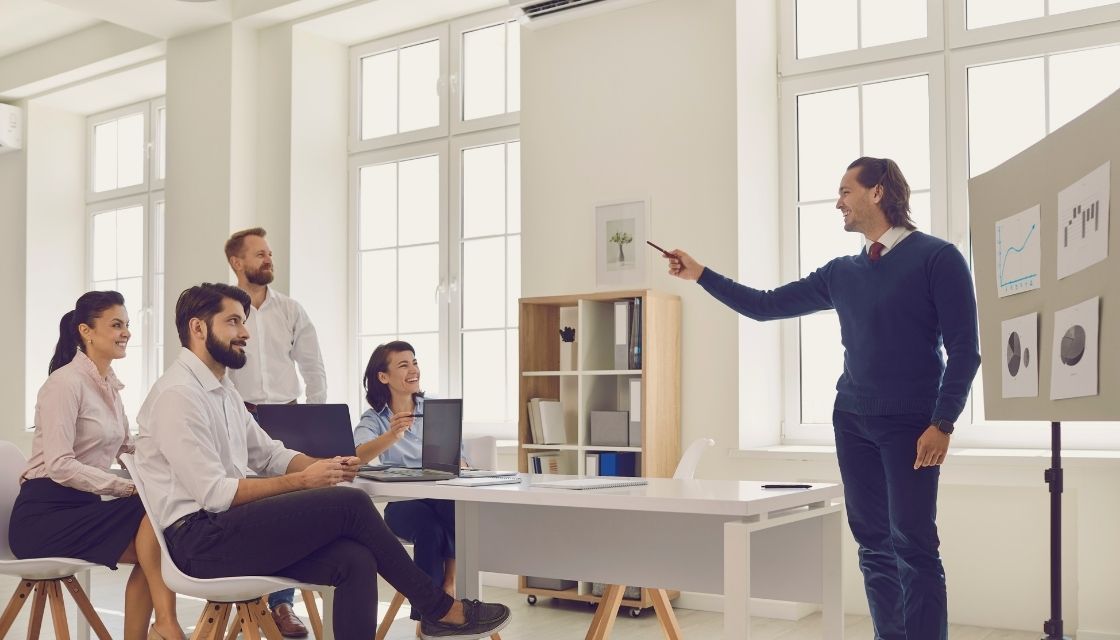 Ein Meeting in einem hellen, modernen Büro, bei dem alle Teammitglieder lachen und ein engagierter Präsentierender mit humorvoller Gestik auf sie eingeht. Dieses Bild zeigt, wie Humor in der Führung die Produktivität steigert und Führungskompetenz auf eine sympathische und zugängliche Art präsentiert. Hier wird deutlich, wie positiv sich Humor auf die Teamdynamik auswirkt.