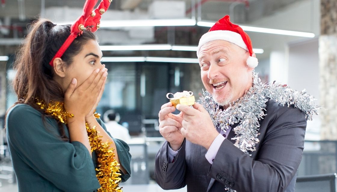 Ein älterer Chef trägt Weihnachtsaccessoires, darunter ein Rentiergeweih und glitzernden Christbaumschmuck, während er einer überraschten Kollegin ein Geschenk überreicht. Dieses Bild symbolisiert, wie Humor in der Führung die Arbeitsatmosphäre lockern und die Zusammenarbeit in Teams stärken kann. Solche Führungskompetenzen schaffen eine vertrauensvolle und inspirierende Unternehmenskultur.