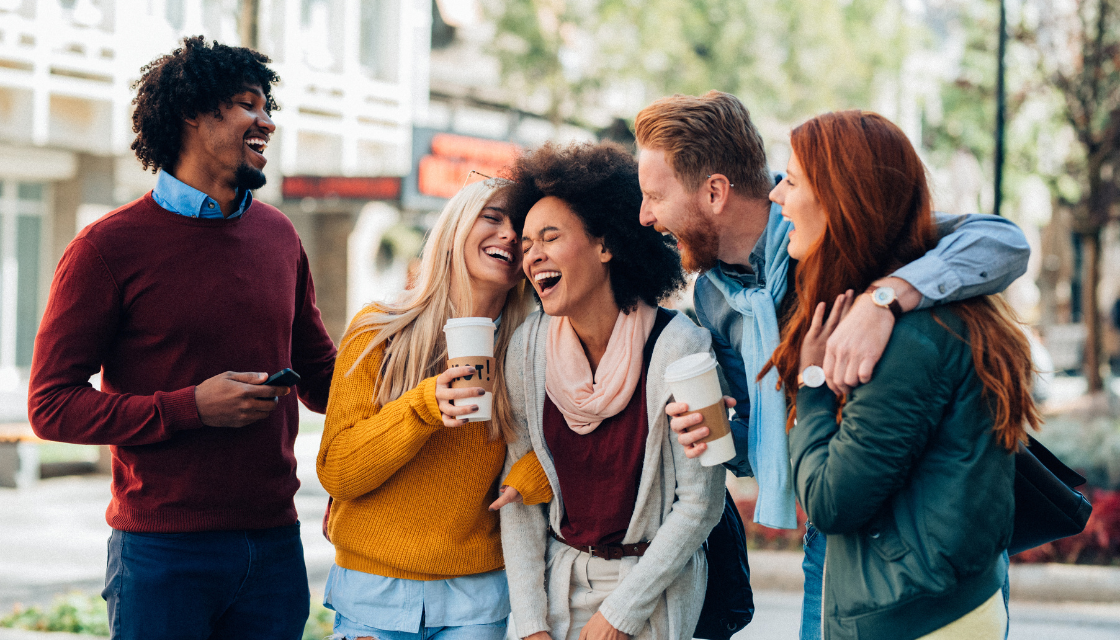 Eine Gruppe von fünf Freunden lacht gemeinsam in der Stadt, während sie Kaffee trinken. Sie umarmen sich und strahlen Lebensfreude aus. Lachen ist gesund – gemeinsam lachen stärkt die Freundschaft und das Wohlbefinden.