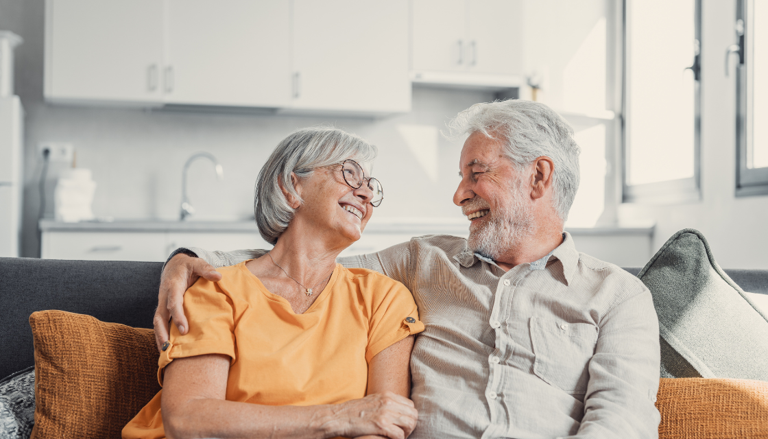 Ein älteres Ehepaar sitzt lachend auf dem Sofa in einer modernen Küche. Beide wirken entspannt und glücklich. Lachen ist gesund – es stärkt die Verbindung und sorgt für Lebensfreude in jedem Alter.