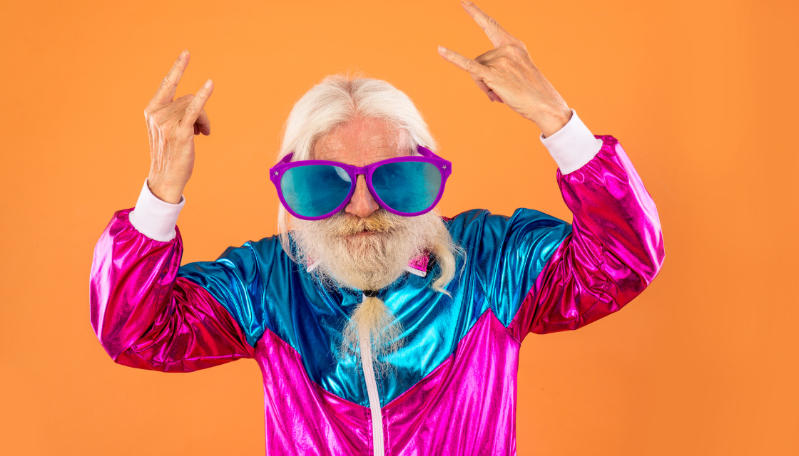 Ein älterer Mann trägt ein buntes, glänzendes Party-Outfit und posiert mit übergrossen Sonnenbrillen in einer rockigen Geste. Das Bild zeigt, wie Humor im Alter hilft, konventionelle Rollen zu durchbrechen und die eigene Persönlichkeit frei auszuleben. Die energievolle Szene vermittelt Mut, Spass und die Botschaft, dass Alter keine Grenzen für Humor kennt.