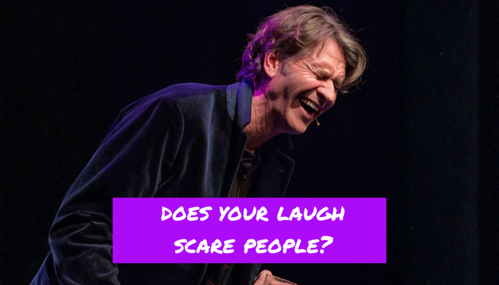 Rob Spence lacht auf einer Bühne unter der provokanten Frage „Does your laugh scare people?“. Das Bild greift humorvoll das Thema auf, warum Menschen ihr Lachen verbessern möchten.